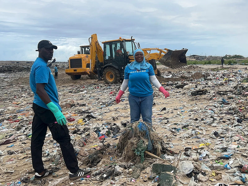 Two people standing in a field of plastic waste with a yellow machine behind them