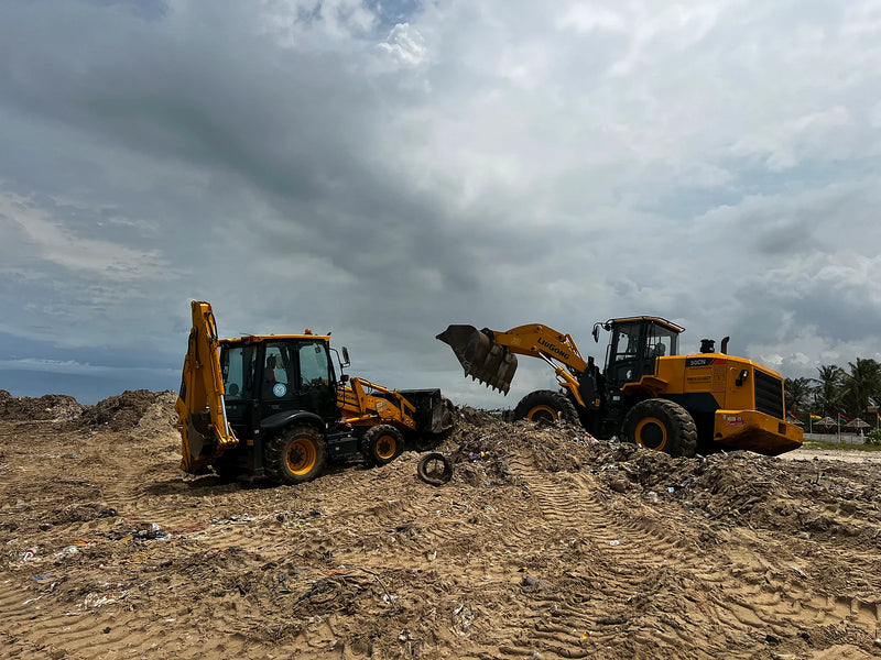 Two yellow machines standing beside a debris mound on sandy ground