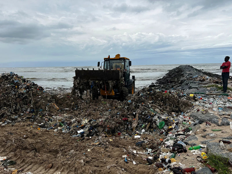 Yellow loader lifting a bucket of waste from a beach covered in plastic bottles