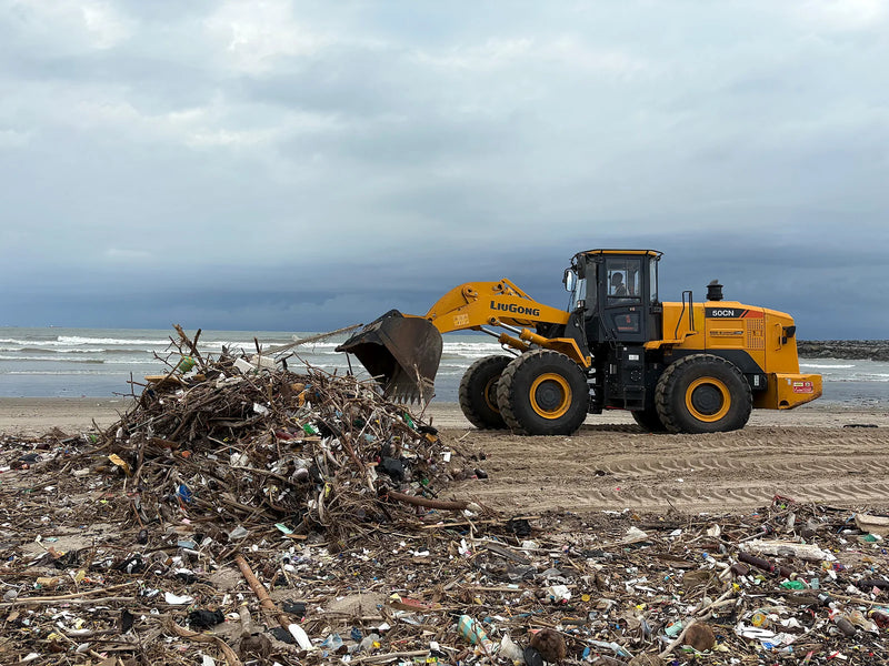 Yellow loader facing a large pile of driftwood and plastic on a beach