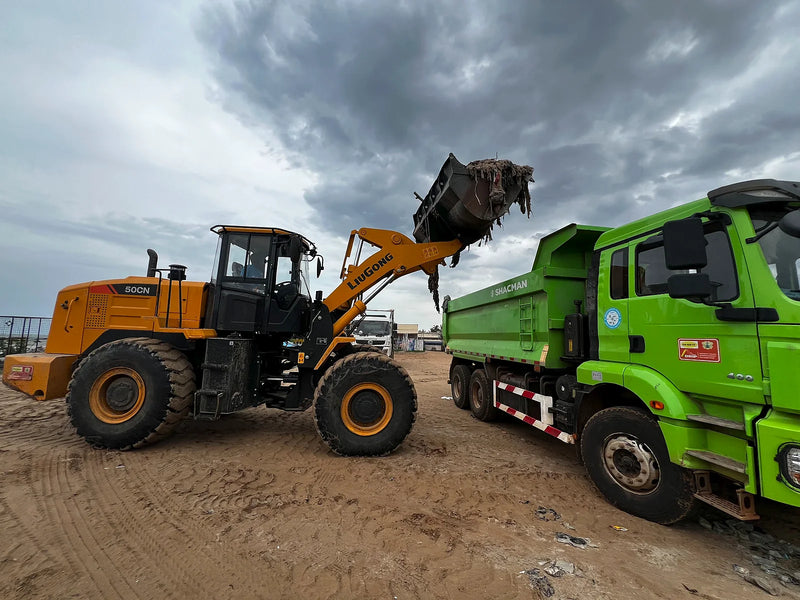 Yellow loader emptying debris into a green dump truck on sand