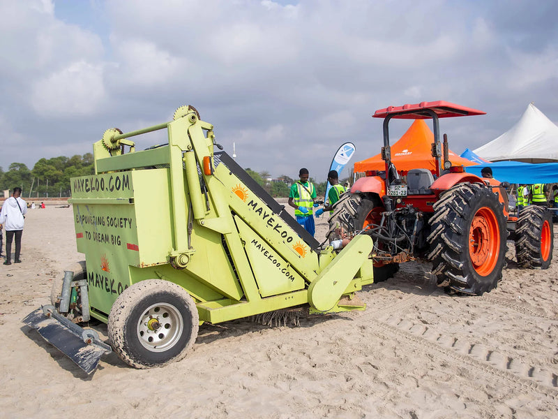 Green beach-cleaning machine hitched to a red tractor on sand near tents
