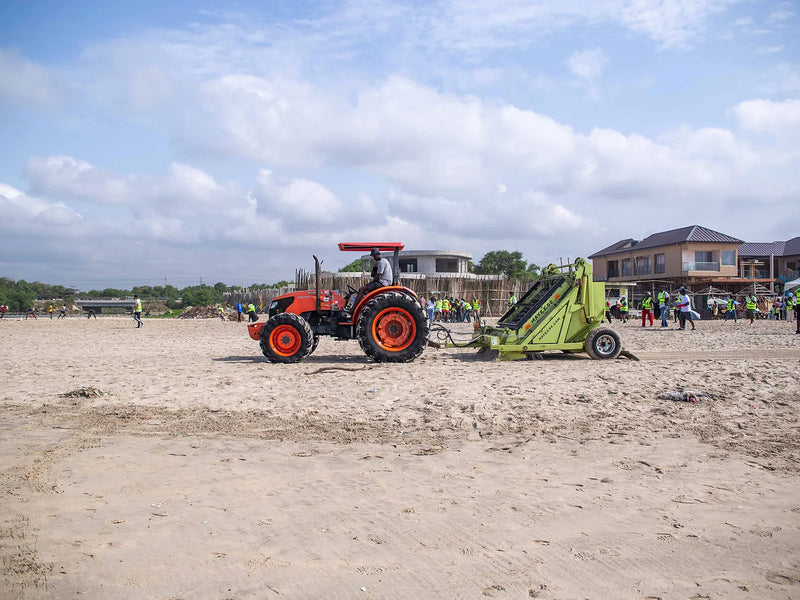 Orange tractor towing a green beach-rake machine across sand